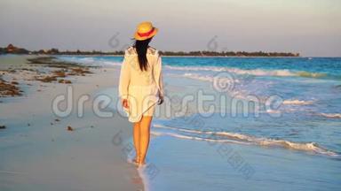日落时热带海滨的年轻美女。 晚上在海滩上穿裙子的快乐女孩。 慢速视频